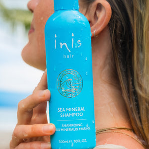 A woman outdoors holds a bright blue bottle of inis hair Sea Mineral Shampoo against her shoulder. The bottle is wet with water droplets, and the background shows a blurred beach scene.